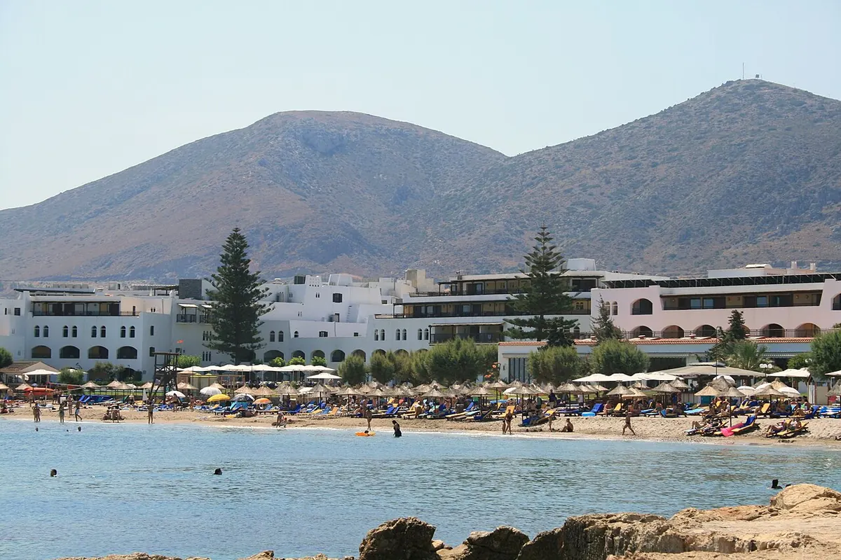 A sandy resort beach with sunshades below white hotel buildings and a brown mountain behind (the beach at Hersonissos on Crete's north coast, not the theatre)