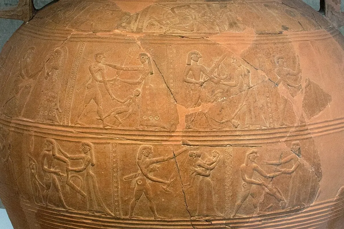 A large clay storage jar with bands of relief figures, displayed against a concrete wall (the Mykonos vase of about 675 BC, which shows the sack of Troy and the wooden horse)